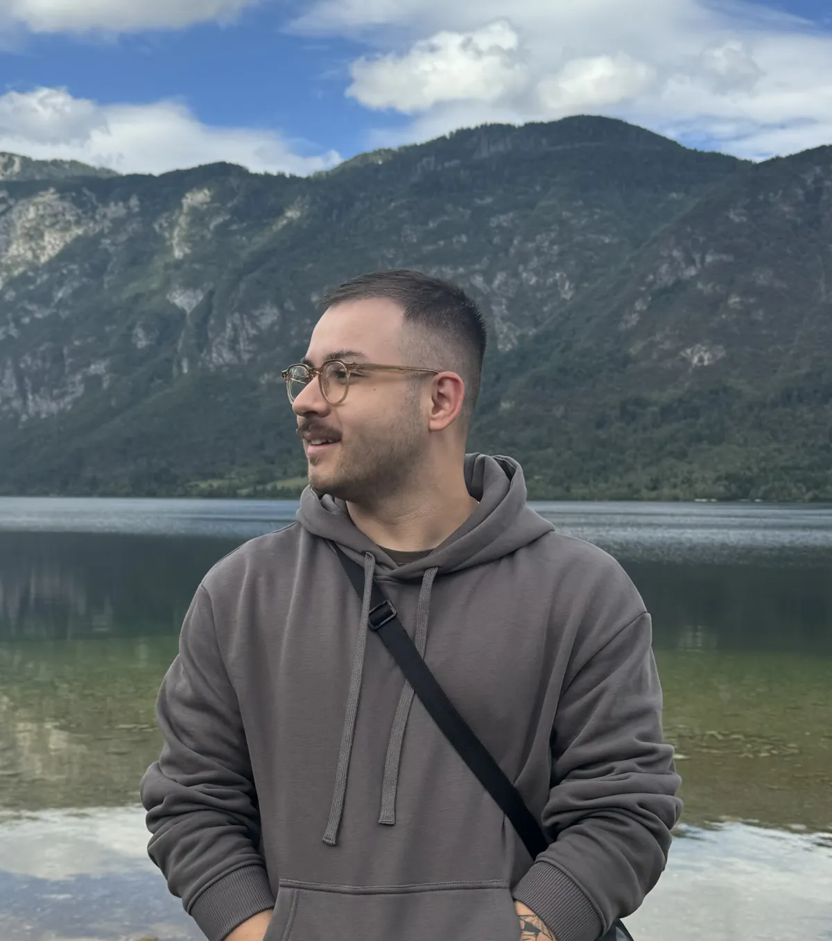 Nícolas de Queiróz, wearing glasses and a hoodie, by a lake among mountains
