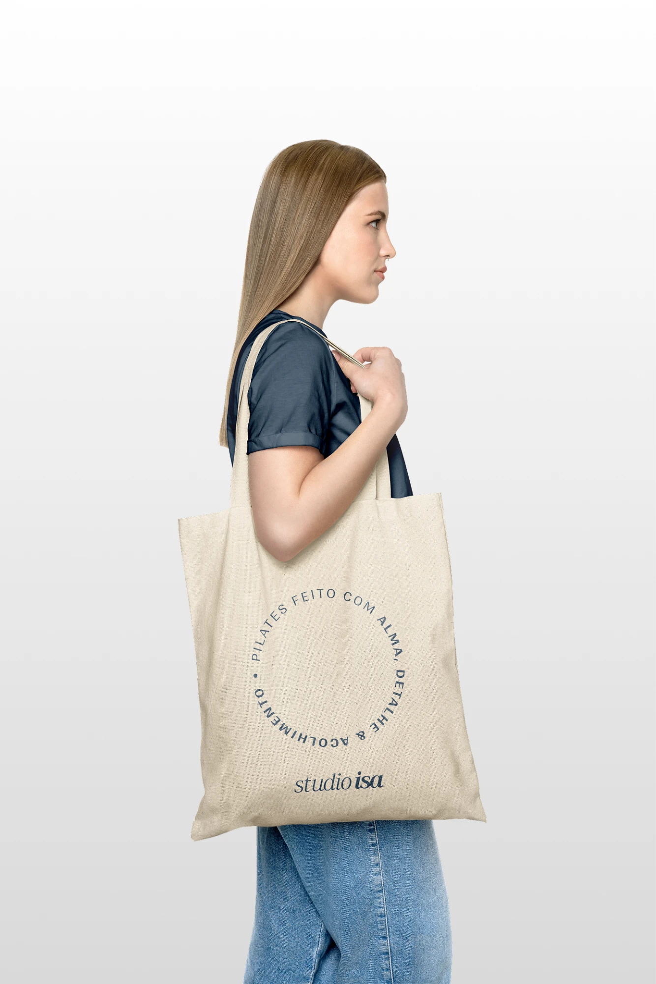Woman in profile holding a raw cotton tote bag with the circular seal Pilates feito com alma, detalhe & acolhimento and the studio isa logotype in blue
