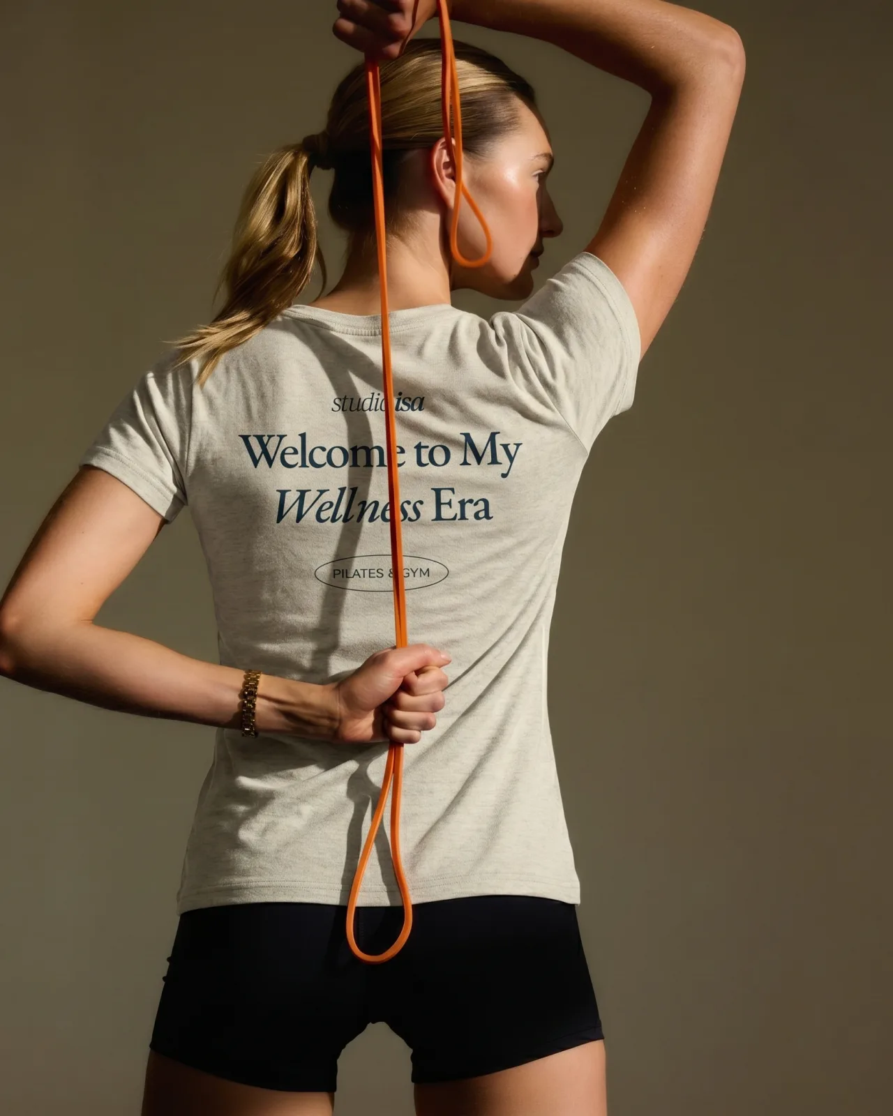 Woman seen from behind with an orange resistance band, wearing the beige Welcome to My Wellness Era T-shirt