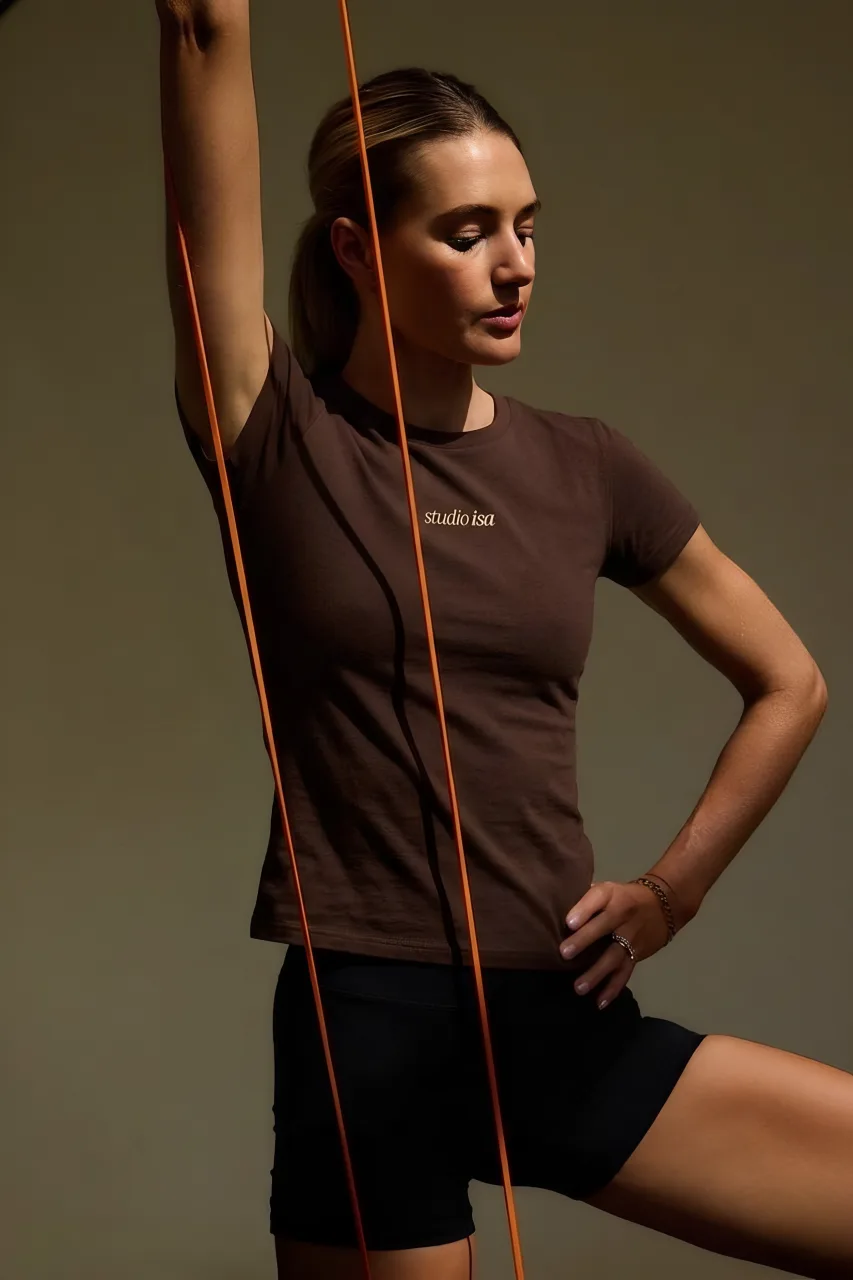 Woman standing with a hand on her hip and an orange band, wearing a brown T-shirt with the studio isa logotype on the front