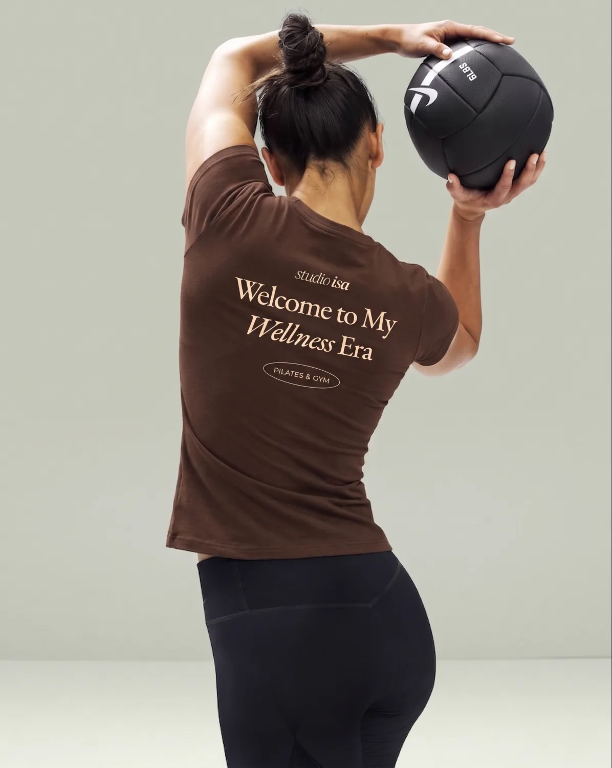 Woman seen from behind holding a medicine ball overhead, wearing the brown Welcome to My Wellness Era T-shirt