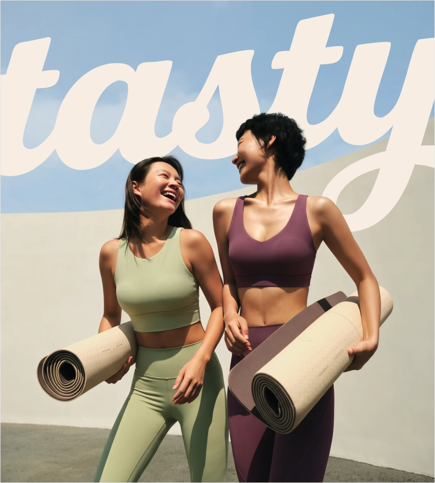 Two women laughing with yoga mats beneath the cream tasty lettering on a wall with a blue sky